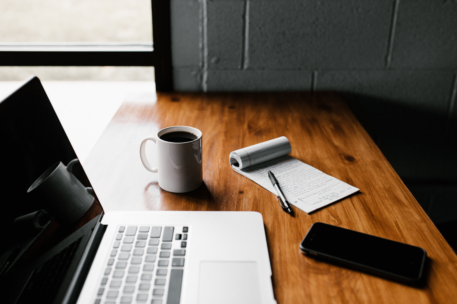 A table with coffee mug, pen, notepad , phone and laptop on it