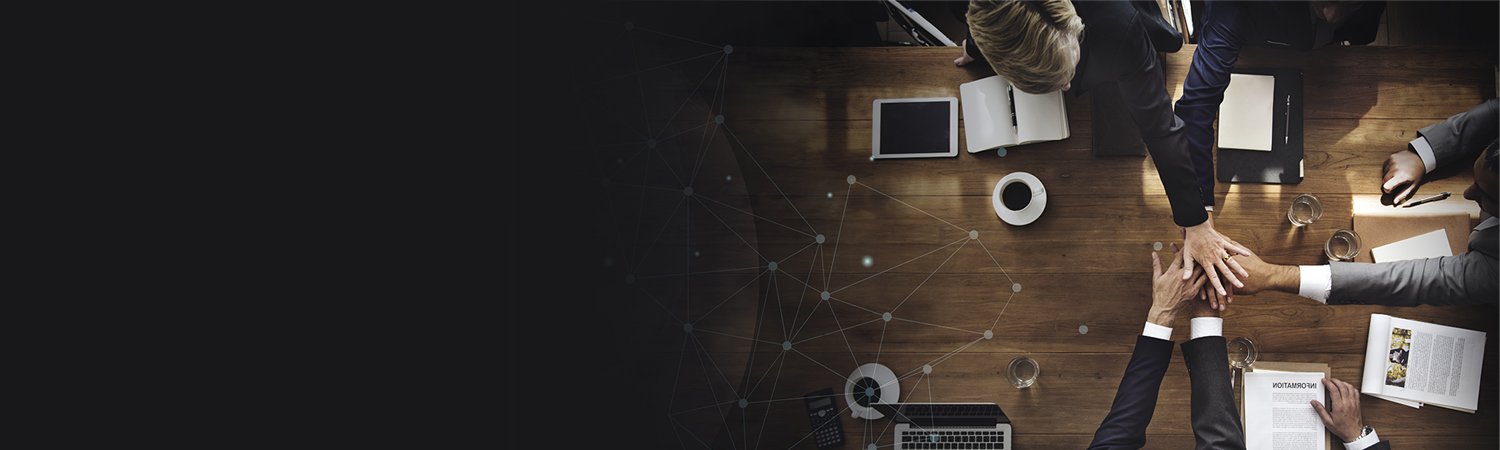 A top-down view of a group of business people joining hands over a desk with Teams of Use laptops, papers, and a cup of coffee, symbolizing teamwork and collaboration in a professional setting.