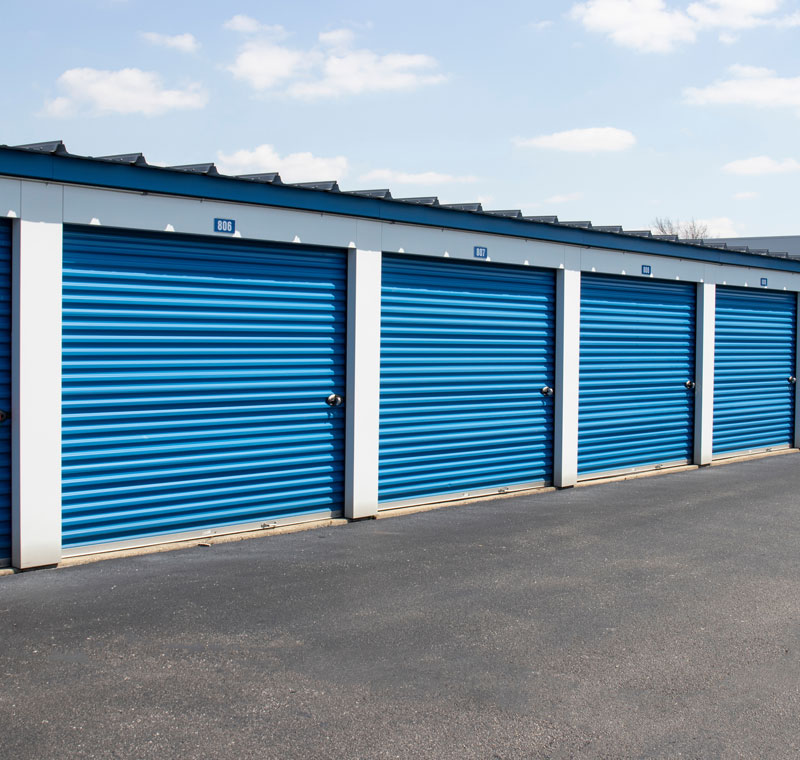 A row of blue self-storage units with metal roll-up doors. The units are numbered (806, 807, etc.) and set against a clear sky with clouds. The ground is paved with asphalt.
