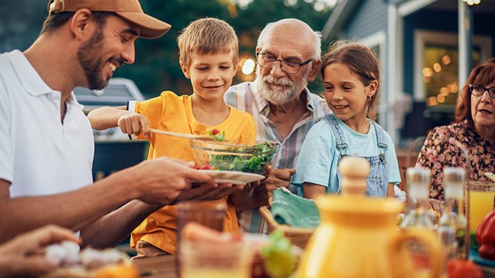 A family enjoying a meal together outdoors. A man is serving a bowl of salad to two children, a boy and a girl, while an elderly man and a woman smile nearby. The scene is warm and joyful with a variety of food and drinks on the table.