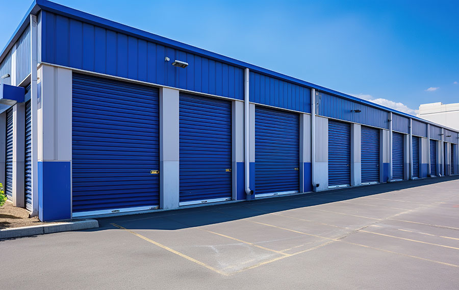 Close-up view of blue self-storage roll-up doors, lined up in a row, with a clear sky in the background and a light fixture above each door. The doors are uniform in size and well-maintained."