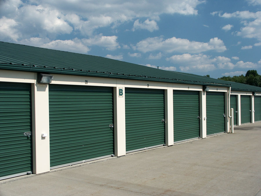 "Exterior view of a self-storage facility with green metal roll-up doors and clear blue skies in the background. The units are numbered, and the facility appears well-maintained."