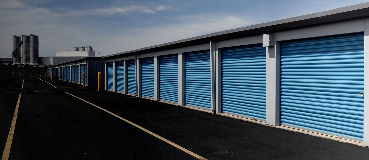 Close-up view of blue self-storage roll-up doors, lined up in a row, with a clear sky in the background and a light fixture above each door. The doors are uniform in size and well-maintained."