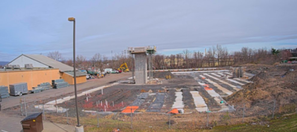 A construction site with a partially constructed building in the background. The ground is being prepared with foundations and rows of material marked with orange. Various construction machinery and equipment, such as a crane and a backhoe, are visible. The sky is cloudy and overcast.