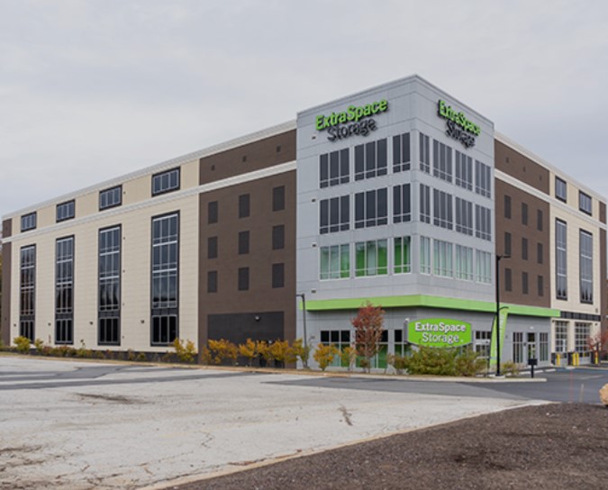 A modern, multi-story ExtraSpace Storage facility with large windows and the company's branding in green on the exterior. The building is located in an open parking area with freshly landscaped surroundings and cloudy skies above.