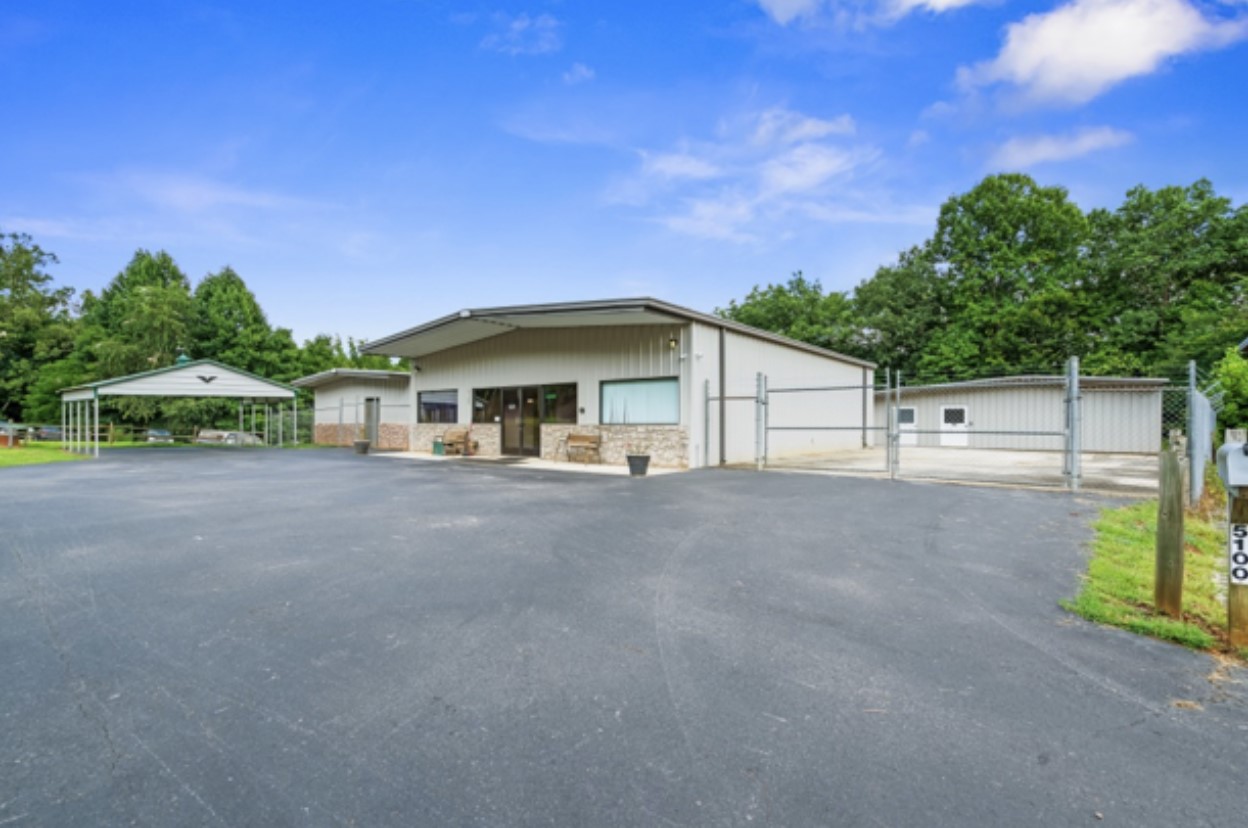 A commercial property with a large paved driveway, a metal building featuring stone accents, and a gated perimeter. The background includes trees and a bright blue sky.