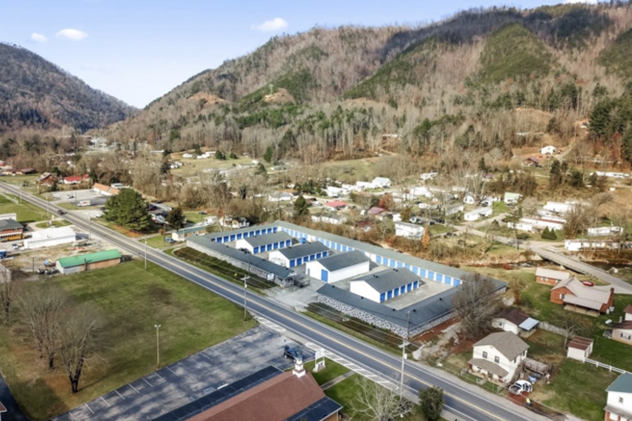 An aerial view of a large industrial construction site, featuring a partially built structure with a metal frame and concrete walls. Construction equipment, workers, and a crane are visible on the surrounding dirt ground.