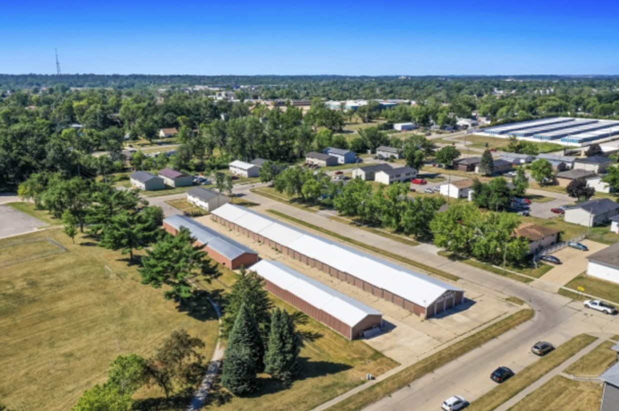 An aerial view of a large industrial construction site, featuring a partially built structure with a metal frame and concrete walls. Construction equipment, workers, and a crane are visible on the surrounding dirt ground.