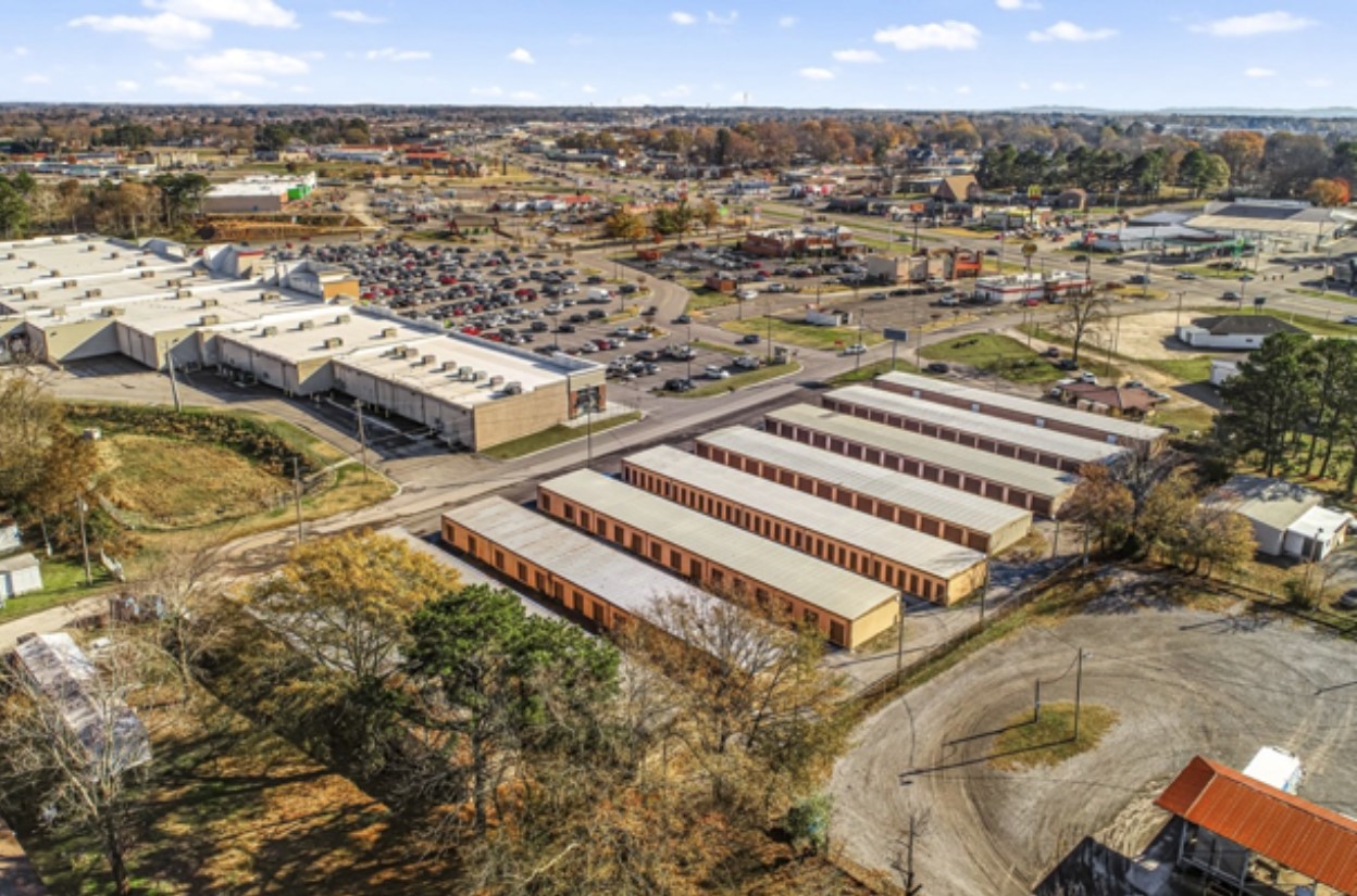 An aerial view of a large industrial construction site, featuring a partially built structure with a metal frame and concrete walls. Construction equipment, workers, and a crane are visible on the surrounding dirt ground.