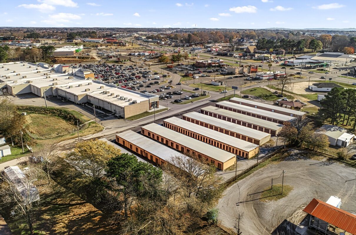 Aerial view of a self-storage facility with multiple storage unit buildings in the foreground, surrounded by roads, parking lots, and commercial buildings in the background. The image shows a broad area with clear views of nearby infrastructure and businesses.
