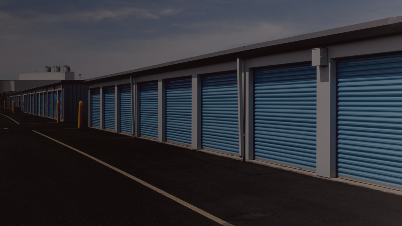 Wide view of a row of self-storage units with blue roll-up doors, arranged neatly along a paved road with yellow bollards. The image highlights the organized, secure nature of the facility.